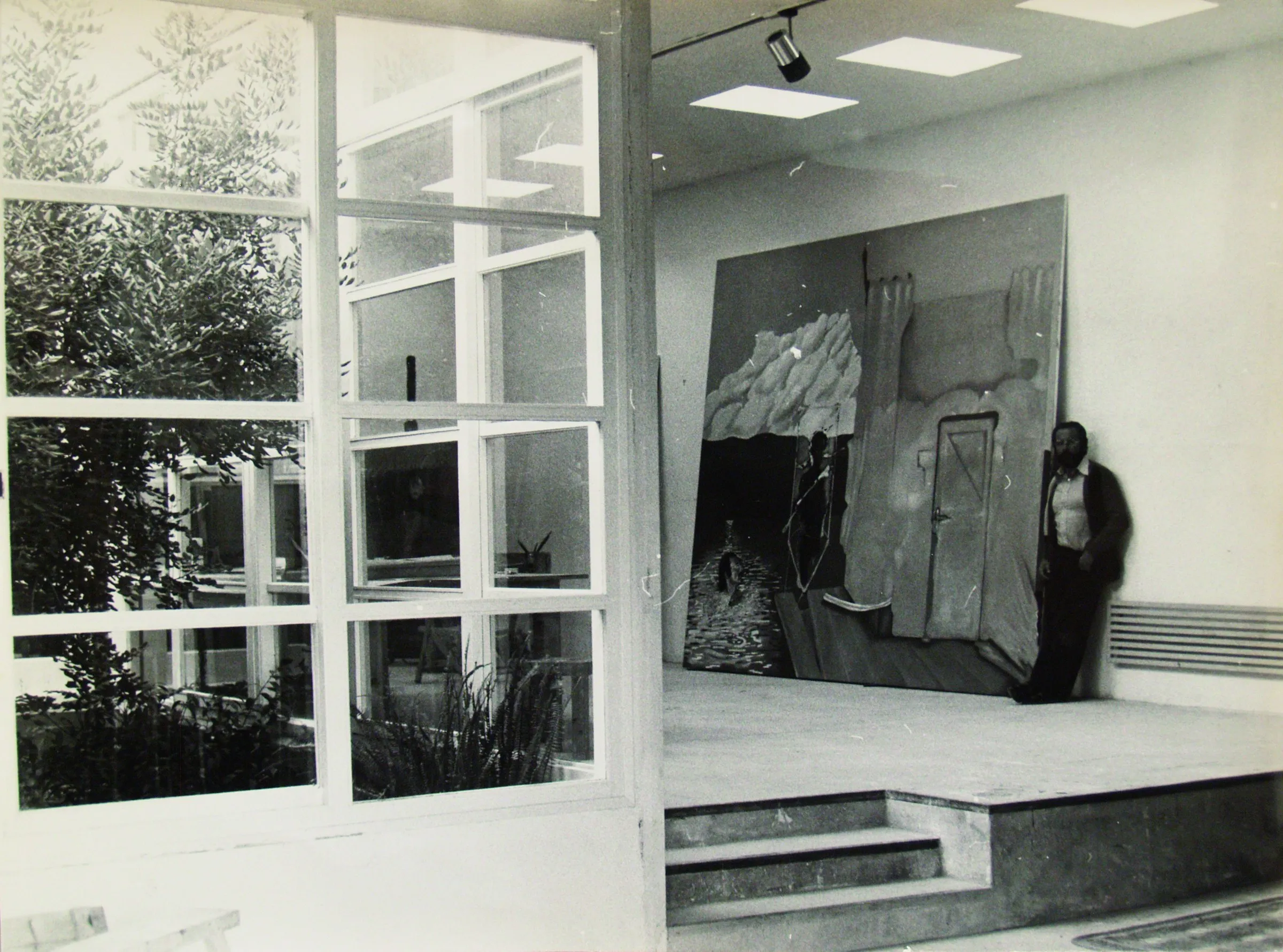 Sala amb una paret de vidre sobre un jardí amb una olivera, probablement a Tarragona. Damunt d'una tarima, una pintura de gran format: un carrer empedrat en perspectiva i unes mans a la part alta. Ell, recolzat a la paret. Fotografia en blanc i negre.