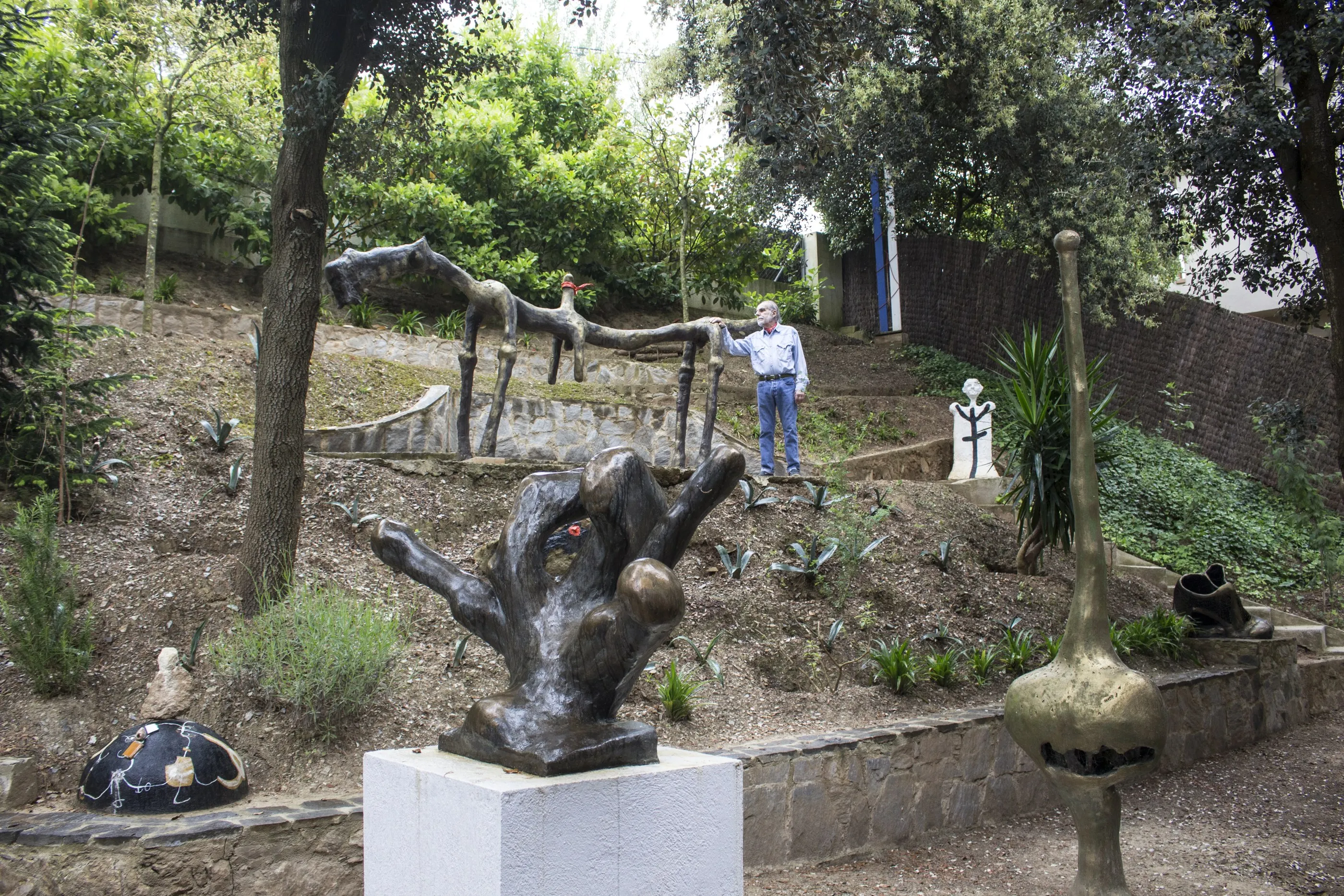 Jardí en pendent amb terrasses de pedra seca. Diverses escultures de bronze a l'aire lliure: una peça horitzontal allargada damunt de potes primes a la terrassa alta, una gran forma de mà damunt d'un pedestal blanc en primer terme, i una forma bulbosa alta a la dreta. Una persona amb camisa blava i cabell blanc és dreta a la terrassa alta, tocant una de les potes.