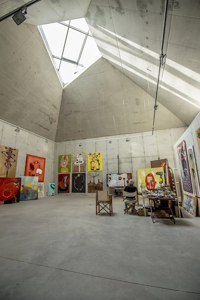 La sala de pintura vista des del terra. Quatre plans inclinats de formigó vist pugen des de les parets i es troben en un lluernari de vidre, un quadrilàter irregular pel qual entra tota la llum. A baix, algú assegut d'esquena davant d'una taula de treball, envoltat de teles recolzades a les parets.