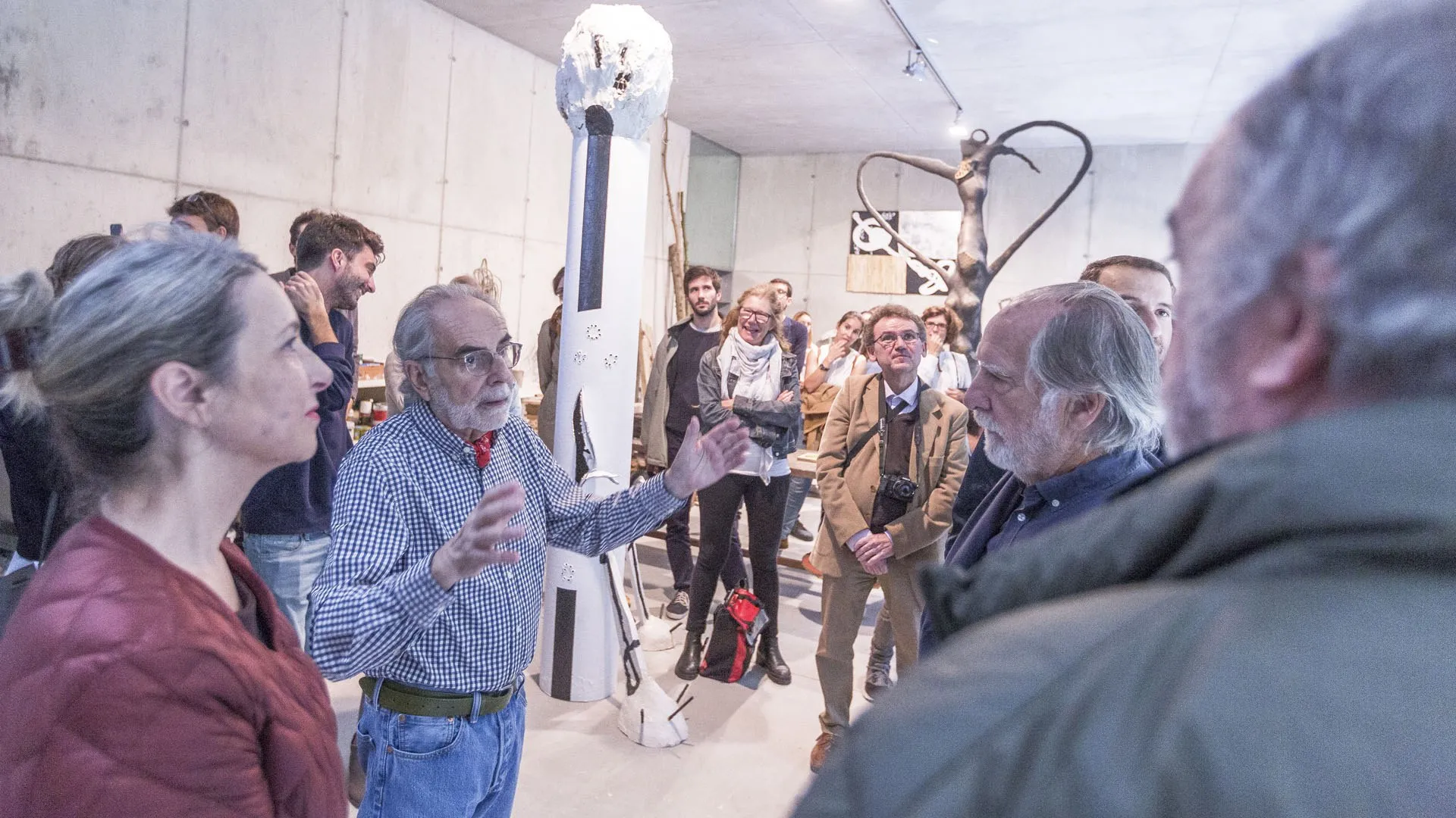 Jornada de portes obertes al taller d'escultura, octubre del 2017: un grup de visitants al voltant d'una persona amb barba blanca que explica, amb les mans obertes, davant de la columna blanca amb el cap de guix.